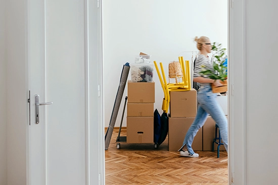 Woman carrying a moving box walking through a hallway with other home goods packed in boxes.